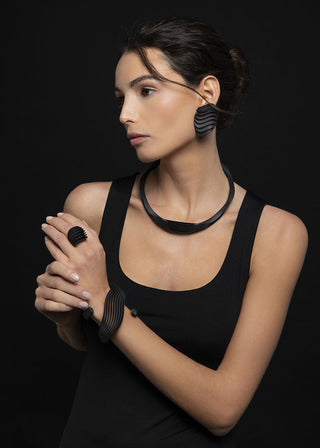 A woman in a black tank top models a matching black jewelry set featuring a thick collar necklace, a wide cuff bracelet, a ring, and large wavy drop earrings against a solid black background.