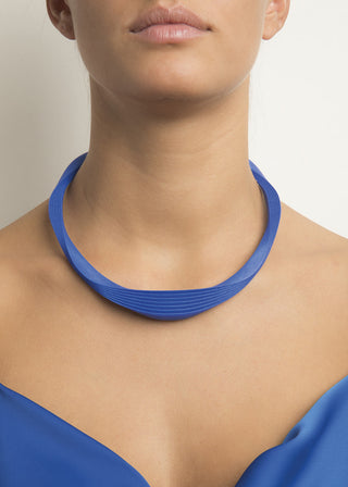 A close-up of a model wearing a wide, sculptural collar necklace in a vibrant royal blue, paired with a matching blue top against a light neutral background.