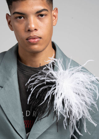 Person wearing a light gray blazer with a feathered accessory on a plain background
