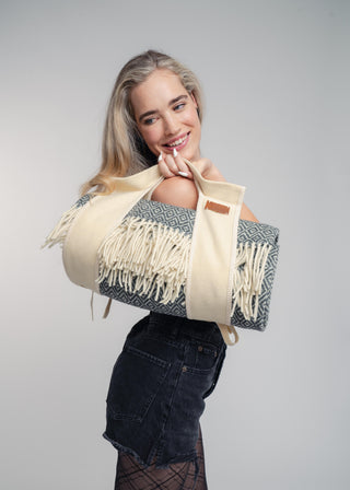 A woman with blonde hair is shown from the waist up, wearing a cream and gray patterned knit wrap sling across her shoulder. She is wearing a dark denim skirt and black tights, posing against a solid light gray studio background.