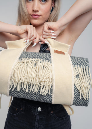 A woman holds a large cream and navy woven tote bag with fringe detailing. The bag features a geometric patterned body, thick cream fabric handles, and a matching cream fringe trim.