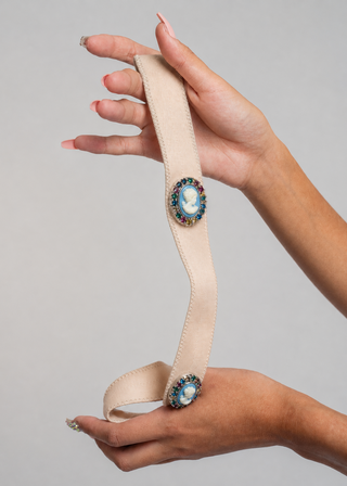 A beige fabric belt with two ornate oval brooch embellishments, held by two hands against a neutral gray background.