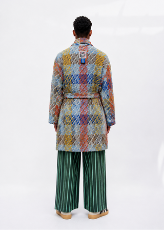 A model viewed from the back wearing a multicolor diamond-patterned quilted coat over a blue and white plaid shirt, paired with green and white vertically striped trousers, against a white studio background.