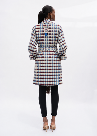A woman viewed from the back wearing a long-sleeve belted coat with a black, white, and red geometric pattern, paired with black pants and beige heels, against a white studio background.