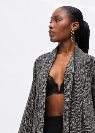 A woman modeling a textured gray open-front cardigan with tassel ties and large patch pockets, paired with black pants and a black bralette, against a clean white studio background.
