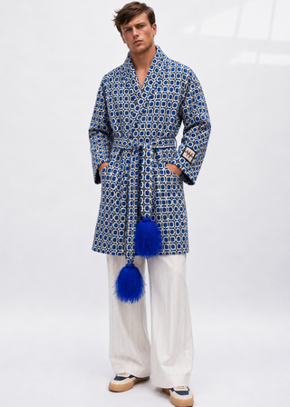 A man modeling a blue and white patterned robe with blue tassels, paired with white trousers and beige sneakers, against a white studio background.