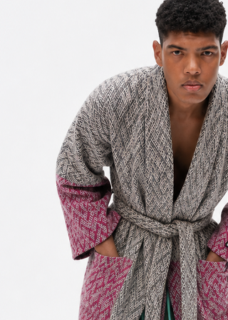 A model wearing a chunky gray cable-knit robe with pink patterned cuffs and hem, green and white striped pants, and pink fluffy tassels, posed against a solid white studio background.