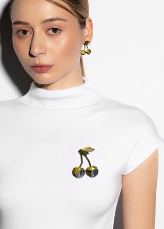 A close-up of a woman wearing a white mock-neck top with a small 3D cherry brooch on the chest, paired with matching cherry-shaped drop earrings.
