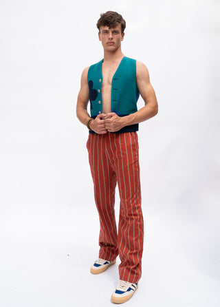 A man modeling a sleeveless teal vest with dark blue circular details, paired with rust-orange pinstripe trousers and white sneakers, against a plain white studio background.