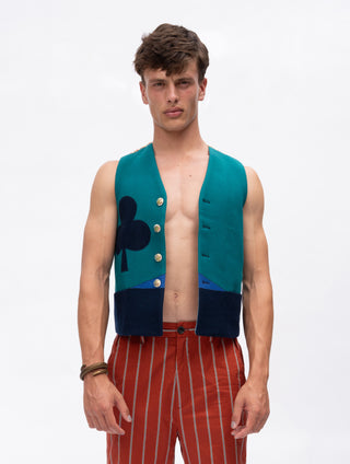 A male model wearing a sleeveless vest with a teal upper section, dark navy lower panels, gold buttons, and a dark navy clover motif, paired with red and white striped trousers, against a plain white studio background.