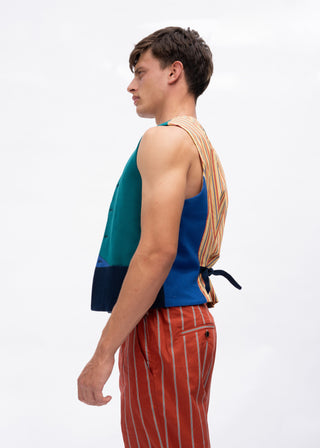 A man shown in profile wearing a sleeveless color-block vest with teal, blue, and navy panels, paired with rust-orange pinstriped trousers. A striped shirt is tied around his back.