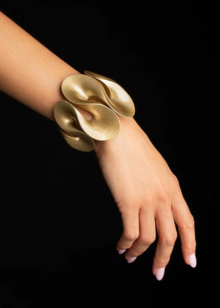 A close-up of a person's wrist and hand wearing a chunky, sculptural gold cuff bracelet against a solid black background.