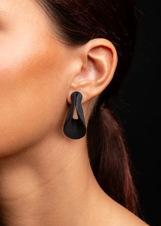 A close-up of a model's ear wearing a matte black teardrop-shaped stud earring with a central cutout, set against a solid black background.
