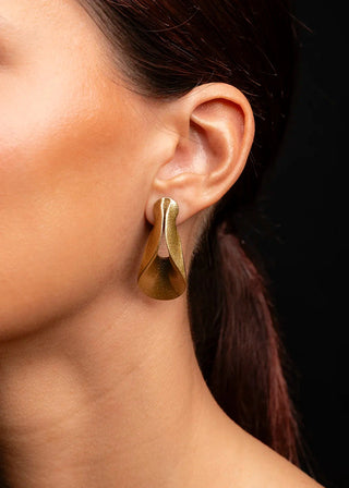 A close-up of a model's ear wearing a single sculptural, organic-shaped gold hoop earring against a solid black background.