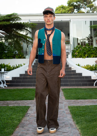 A man walks down a stone path in a landscaped garden, wearing a teal knit vest and tie with dark brown trousers and a cap.