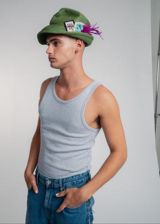 Man wearing a green cap with decorative elements, gray tank top, and blue jeans against a plain background