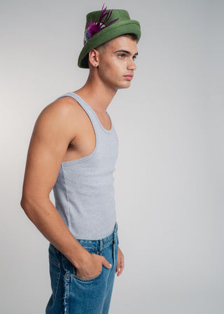 Man wearing a green hat with feathers, gray tank top, and blue jeans on a plain background