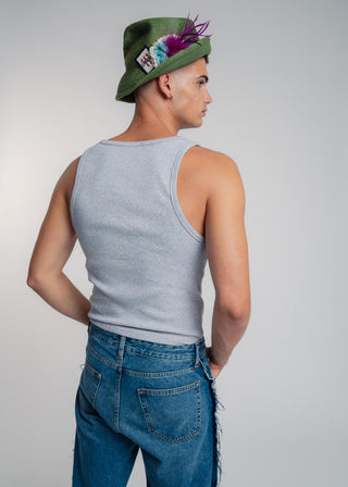 Person wearing a green hat with feathers, gray tank top, and blue jeans on a plain background