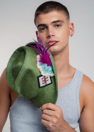 Man holding a green hat with colorful feathers against a plain background