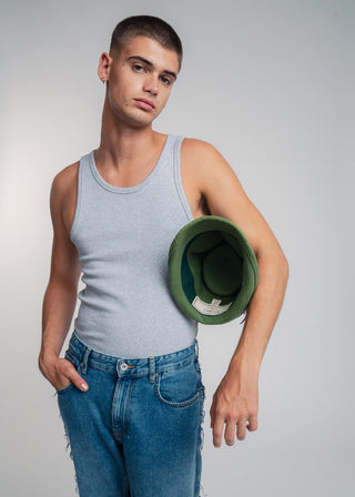 Man wearing a gray tank top and blue jeans holding a green hat against a plain background