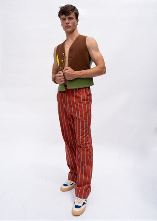 A man modeling a retro-style outfit consisting of a brown vest over a green shirt, paired with rust-red pinstriped trousers and blue-and-white sneakers, against a plain white studio background.
