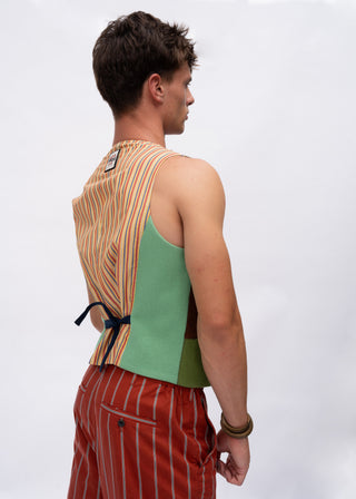 A man shown from the back wearing a sleeveless vest with a striped orange and beige back panel and a solid green side panel, paired with red and white striped trousers, against a light gray studio background.