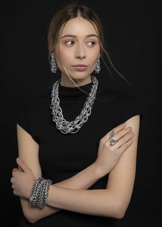 A woman modeling a chunky silver chain-link jewelry set, including a layered necklace, matching drop earrings, and a multi-strand bracelet, against a solid black background.