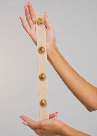 A beige fabric belt with four gold textured buttons, held vertically by two hands against a solid light gray background.