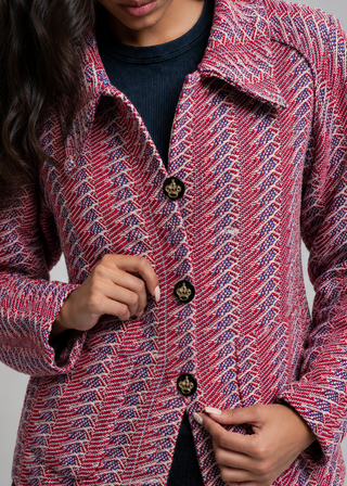 A woman wearing a textured tweed blazer jacket in a red, pink, and blue pattern with gold buttons, paired with a dark navy t-shirt and black jeans, against a solid gray studio background.