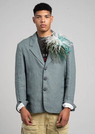 Man wearing a light blue blazer with a feathered accessory on a gray background