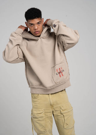 Person wearing a beige hoodie with a logo on the chest pocket against a gray background