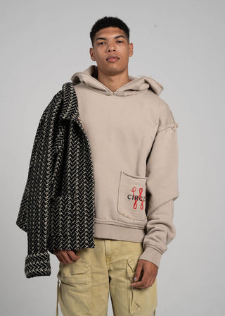 Man holding a patterned coat, wearing a beige hoodie with a logo, on a plain background