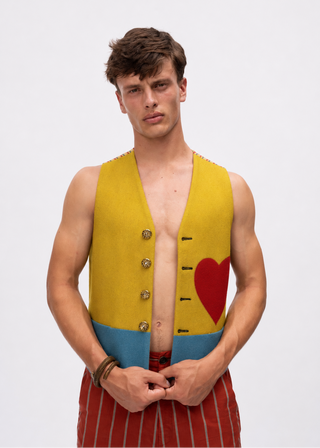 A male model wearing a sleeveless vest with a yellow upper section, blue lower band, gold buttons, and a red heart graphic, paired with red and white striped trousers, against a plain light gray background.
