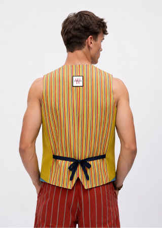 A man viewed from behind wearing a sleeveless yellow vest with vertical red, orange, and blue stripes, tied at the waist with a black ribbon, paired with red pinstriped shorts against a light gray studio background.