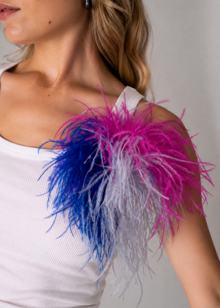 A close-up of a woman wearing a white ribbed tank top and a large, fluffy feathered shoulder accessory in bright pink, blue, and white.