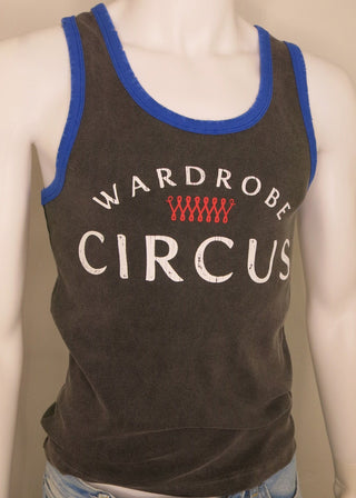 A dark brown tank top with blue trim and white text reading 'WARDROBE CIRCUS' with a red wavy graphic, displayed on a mannequin torso with blue jeans against a plain beige background.
