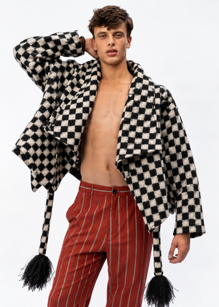 A male model wearing a black and off-white checkered open-front jacket with long tassels and red pinstriped trousers, posed against a plain white studio background.