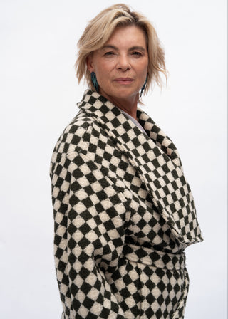 A woman with short blonde hair wearing a black and cream checkered wrap coat, photographed against a plain white studio background.