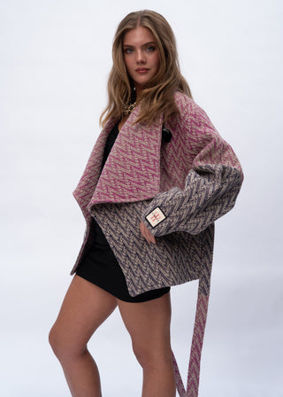 A woman models a pink and gray herringbone patterned wrap jacket with wide sleeves and a long tie belt, styled over a black outfit against a light gray studio background.