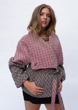 A woman modeling a textured, patterned wrap top with a chevron-like zigzag design in pink, purple, and gray tones, featuring voluminous sleeves and a self-tie belt, paired with black shorts against a plain light gray background.
