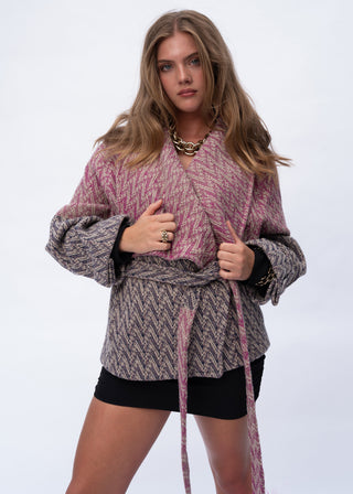 A woman modeling a pink and gray herringbone patterned wrap jacket with a tie waist, styled with black shorts and gold jewelry against a plain white studio background.