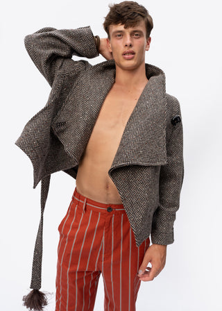A male model wearing a brown and gray herringbone patterned open-front jacket with a tie waist, styled open over a bare torso, paired with red and white vertically striped trousers, against a plain white background.
