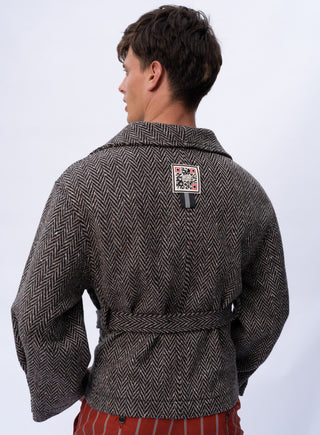 A man shown from the back wearing a dark gray and black herringbone patterned jacket with a matching waist tie belt. The jacket features a small square brand patch on the upper back.