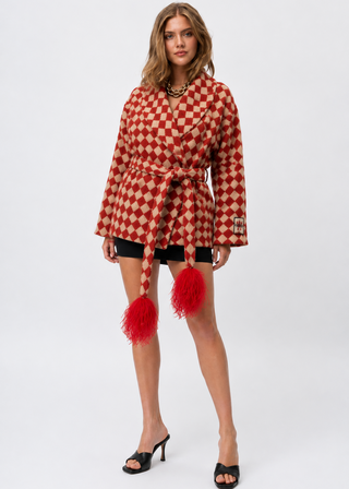 A model wearing a red and white checkered belted wrap coat with red feather trim and tassels, styled with black shorts and black heeled sandals against a light gray studio background.