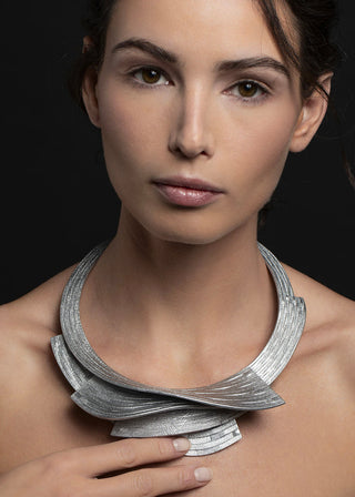 A close-up portrait of a woman wearing a chunky, sculptural silver necklace with layered, ribbon-like bands against a dark background.