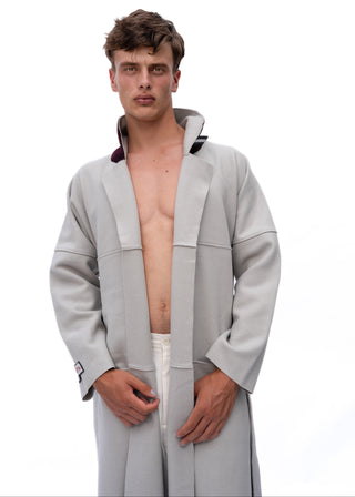 A male model wearing a light gray oversized hooded robe or coat, styled open at the chest, photographed against a plain white studio background.