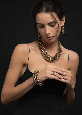 A woman wearing chunky gold-toned jewelry, including a layered chain necklace, matching earrings, and a bracelet, styled with a black spaghetti-strap dress against a dark background.