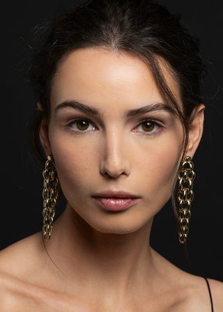 A close-up portrait of a woman with dark hair pulled back, wearing large gold chain drop earrings against a dark black background.
