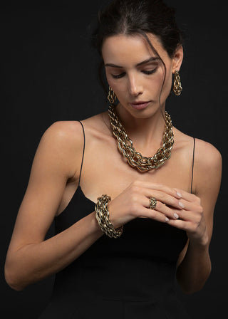 A woman wearing chunky gold-toned jewelry, including a layered chain necklace, matching earrings, and a bracelet, styled with a black spaghetti-strap dress against a dark background.