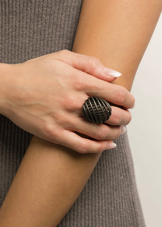 A close-up of a person's hand wearing a large, dark metallic ring with a textured, spherical grid pattern, set against a neutral light background.